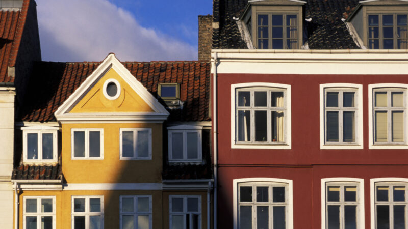 Række af byhuse i gult og mørkerødt med hvide vinduesrammer; det gule hus har en trekantet gavl med et rundt vindue og rødt tegltag, og solen kaster en skarp skygge over den venstre del af facaden mod en blå himmel.
