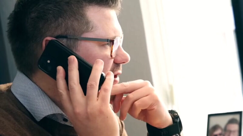 Mand med briller holder en smartphone mod øret og hviler den anden hånd mod hagen, siddende ved et lyst vindue; et sløret indrammet foto af to personer står på et bord i baggrunden.