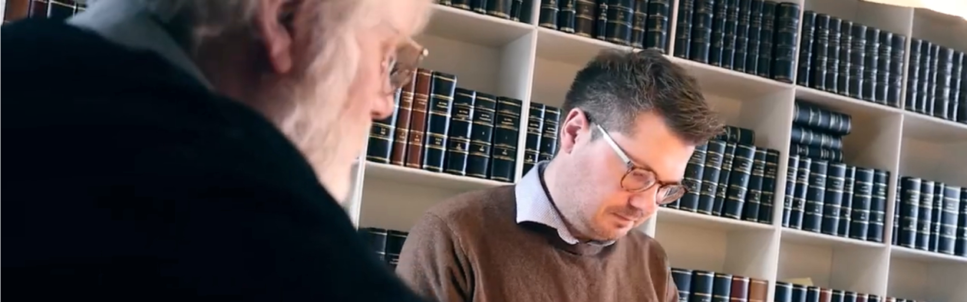 To mænd i et kontor med reoler fyldt med indbundne bøger; en ældre mand med hvidt skæg ses sløret i forgrunden, mens en yngre mand med briller ser ned og læser eller skriver.