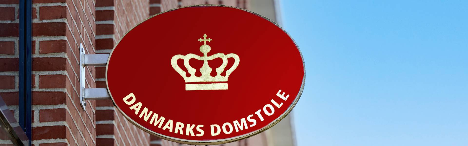 Rødt ovalt skilt med en gylden krone over teksten "DANMARKS DOMSTOLE", monteret på en murstensvæg med blå himmel i baggrunden.