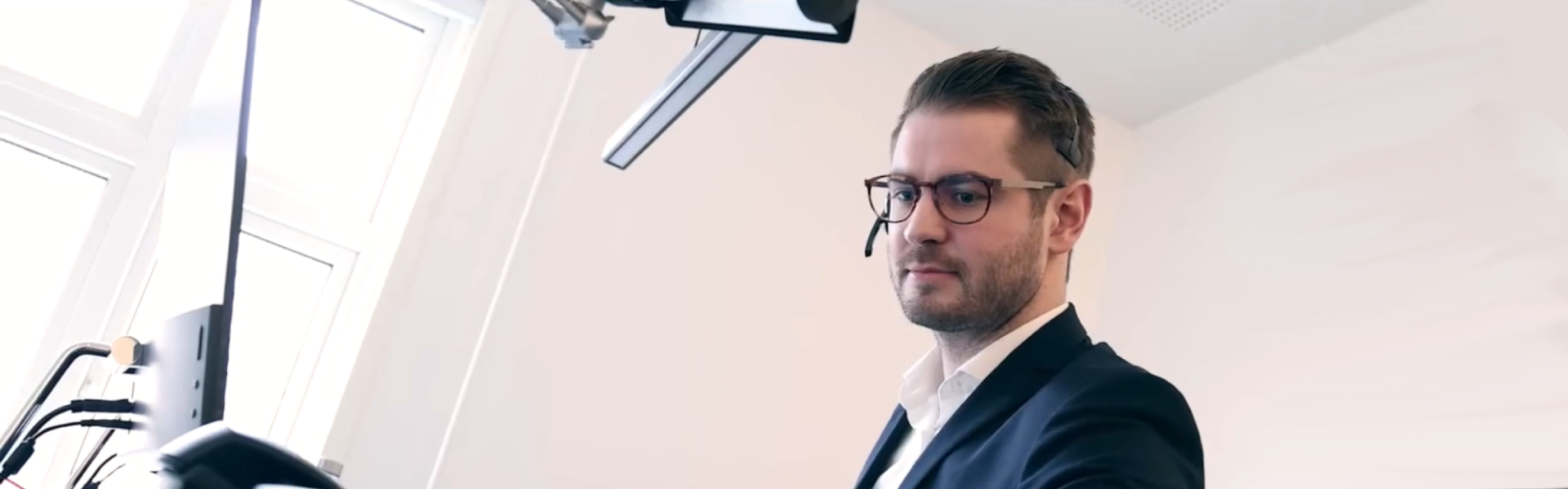 Mand iført jakkesæt, hvid skjorte, brune briller og headset står ved et hæve-sænkebord og kigger på en computerskærm i et lyst kontorlokale med store vinduer.