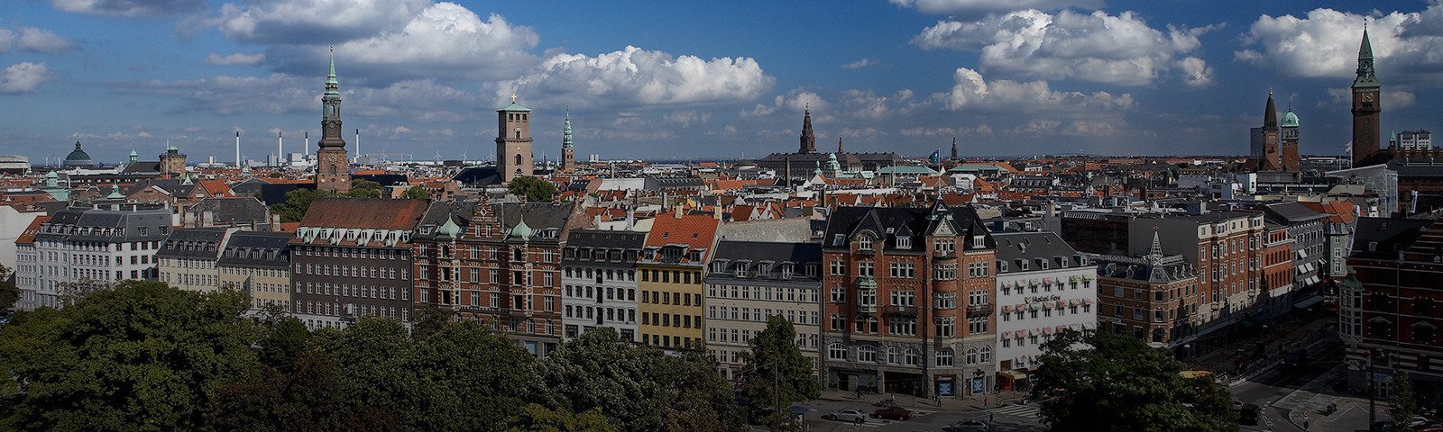 Panorama af København med historiske bygninger, kirketårne og røde tage under en blå himmel med spredte skyer.