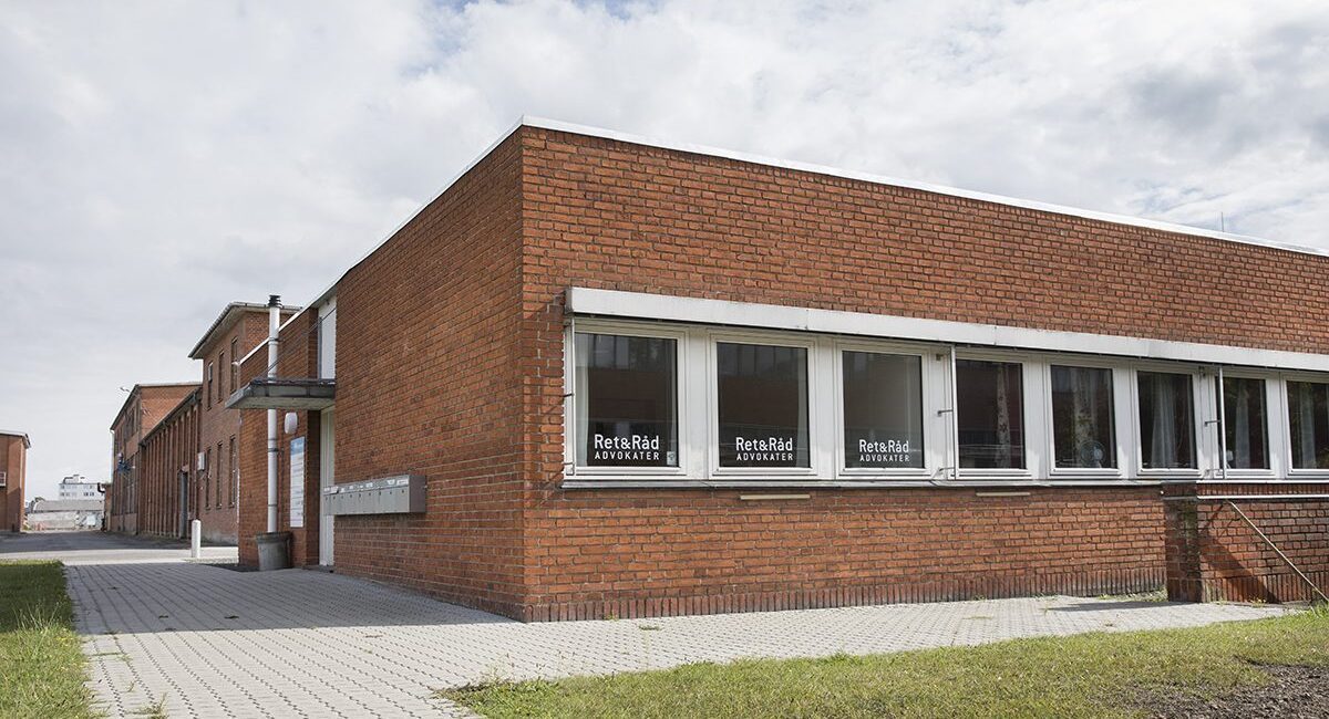 En rødstensbygning med flere vinduer, hvor teksten "Ret&Råd Advokater" ses på glasset. Foran bygningen er der en flisebelagt sti og en græsplæne under en skyet himmel.