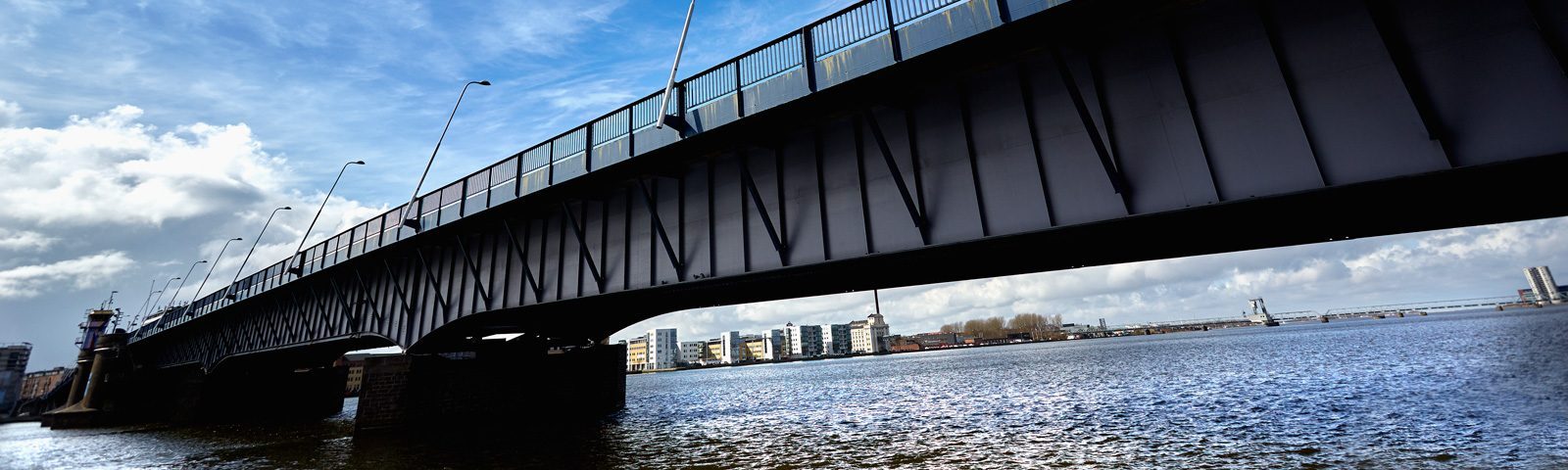 En stor metalbro strækker sig over vandet, med moderne bygninger og en klar blå himmel i baggrunden.